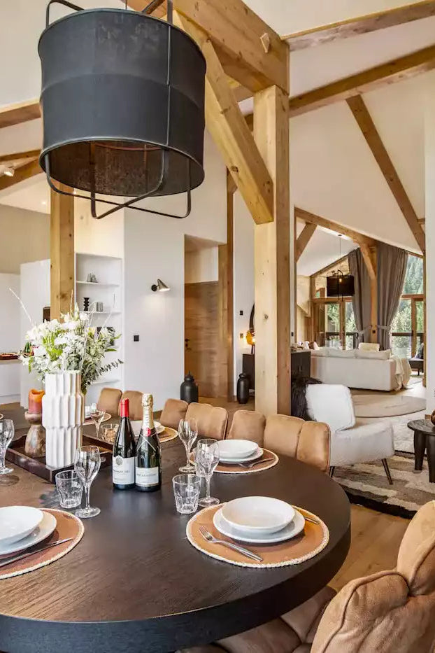 A modern dining room with a dark oval table set for six, surrounded by brown upholstered chairs. The room features exposed wooden beams, a large black pendant light, and an open-plan layout leading to a kitchen and living area.