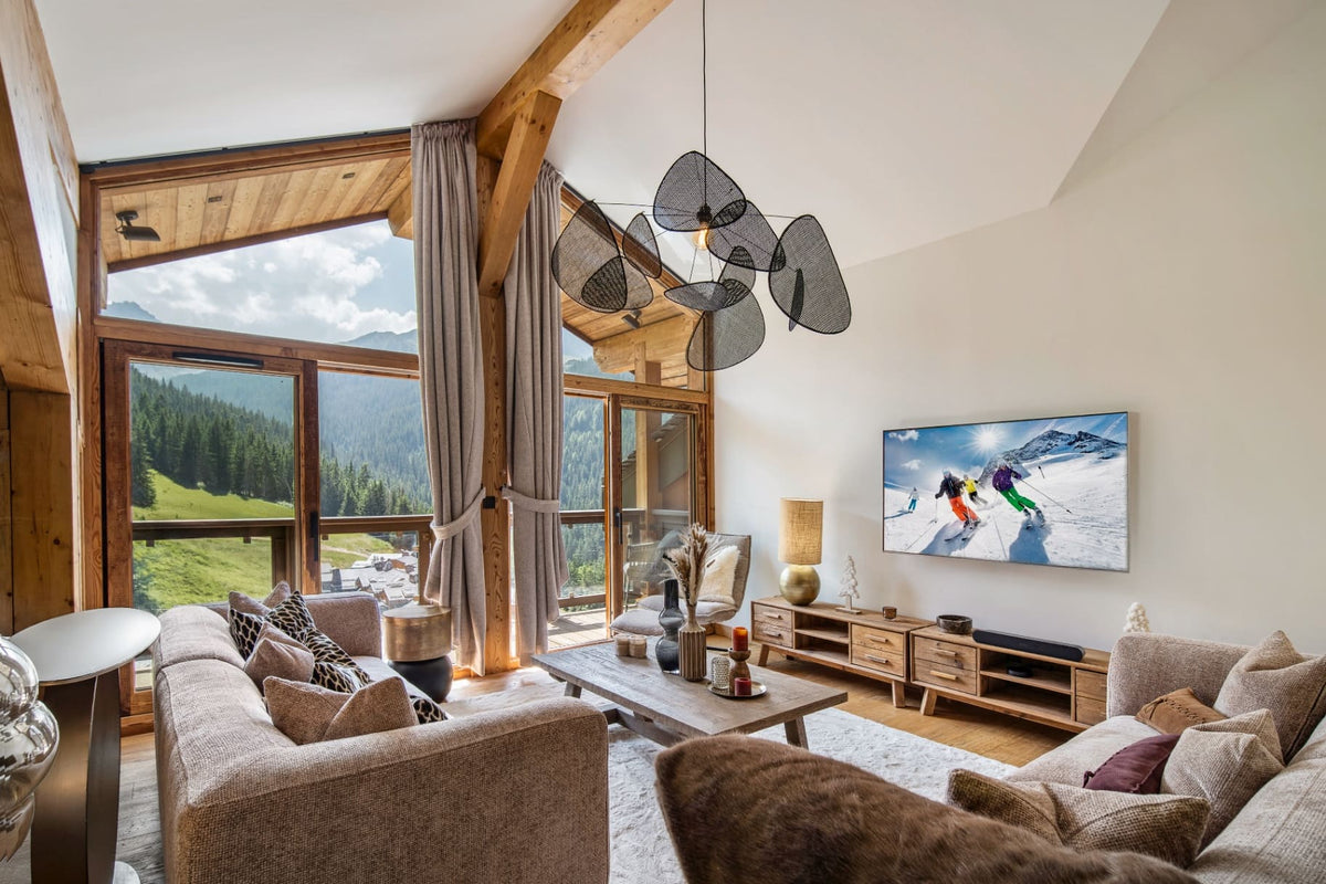 A modern ski chalet living room with a large TV showing a snowy mountain scene, cozy beige sofas, a wooden coffee table, and a distinctive black wire pendant light hanging from exposed wooden beams.