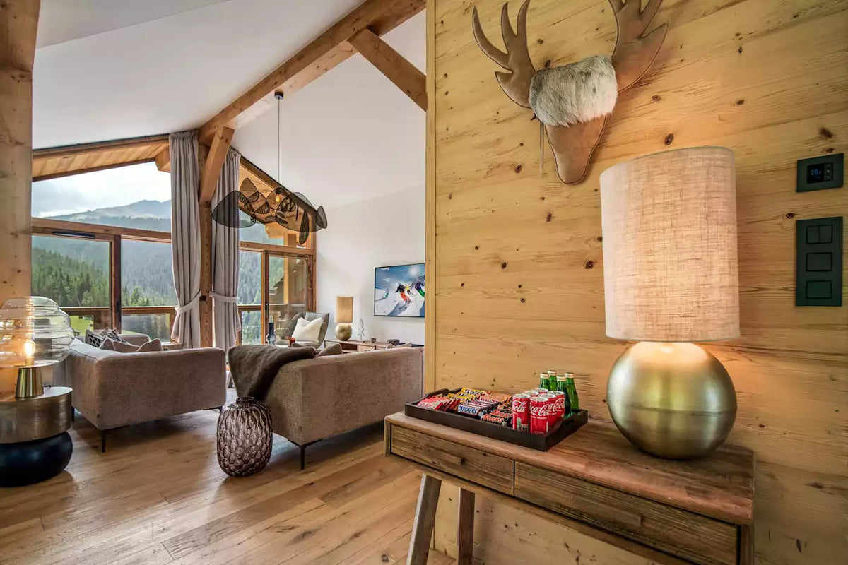 A rustic mountain chalet living room with light wood walls and floors, featuring a glowing table lamp on a wooden console table, a sofa in the background, and a deer head wall mount.