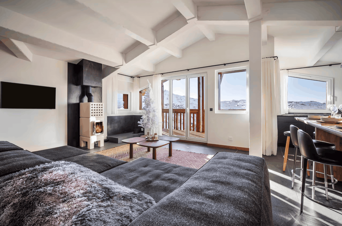 A modern ski chalet living room featuring a large tufted dark gray sofa in the foreground, a black wood-burning fireplace, a small white Christmas tree, and large windows overlooking a snowy mountain landscape.
