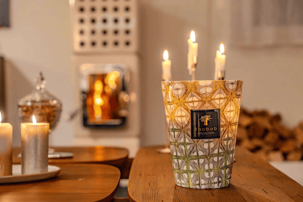A lit candle in a patterned glass holder with three additional candles, styled on a wooden table in a cozy room with a fireplace in the background.