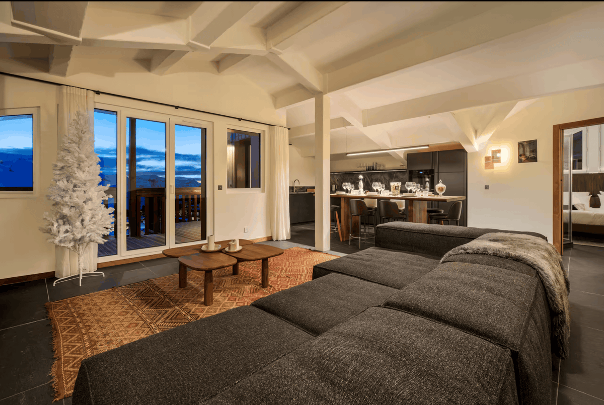 A modern living room interior featuring a large dark gray sectional sofa in the foreground, a white artificial tree near tall windows with a blue sky view, and an open-plan kitchen in the background.