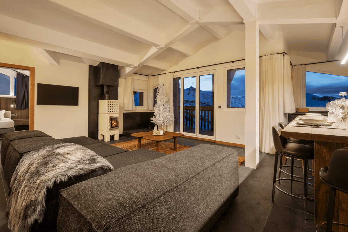A modern ski chalet living room featuring a large dark gray sectional sofa, a white wood-burning stove, a small white Christmas tree, and a wooden dining bar with stools overlooking a mountain view at dusk.