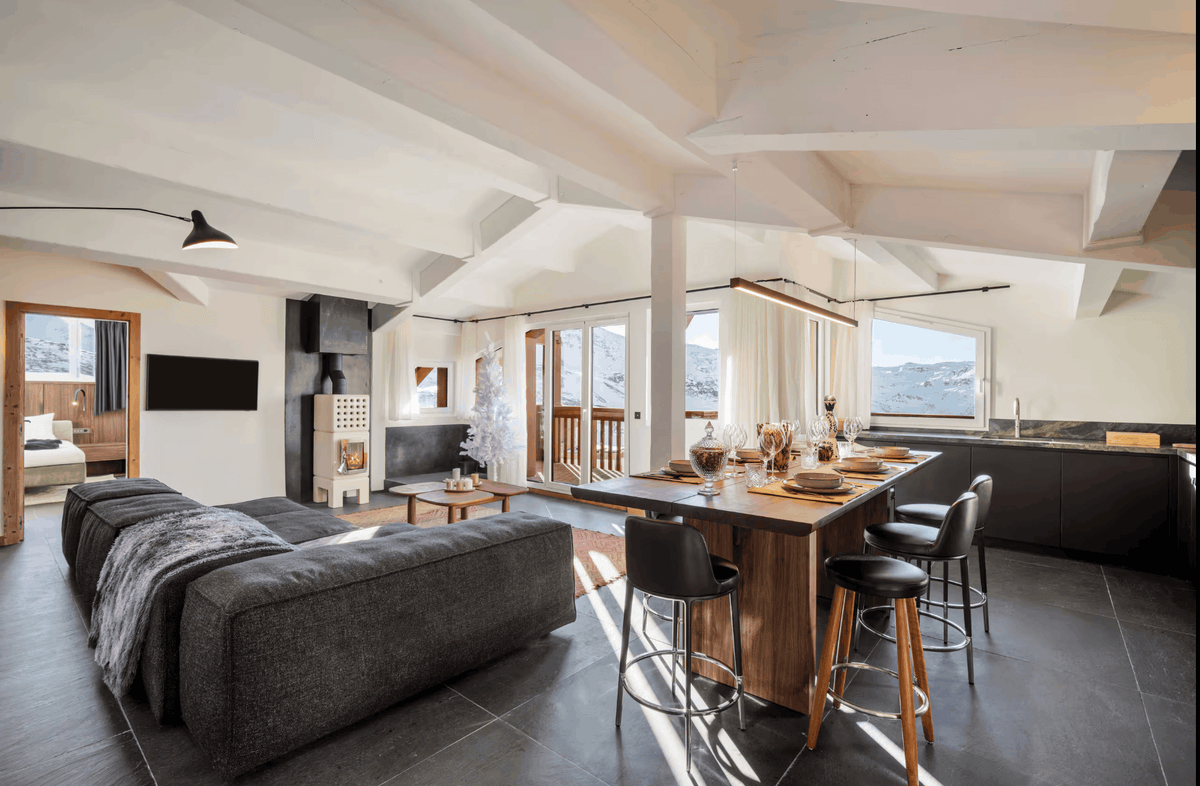 A modern open-plan chalet interior featuring a large dark gray sectional sofa, a wooden kitchen island with three bar stools, a black fireplace, and high ceilings with exposed beams.