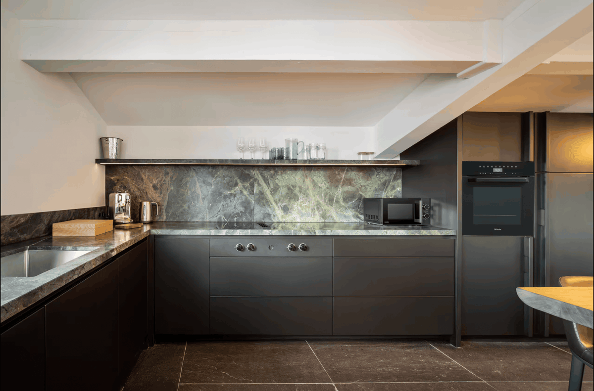 Modern kitchen interior with dark charcoal cabinets, a green-and-white marbled stone backsplash, dark countertops, built-in appliances, and a visible sloped ceiling.