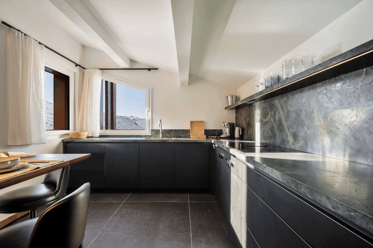 Modern kitchen interior with dark charcoal cabinets, a matching dark stone backsplash and countertop, large windows with white curtains, and a light gray slatted ceiling.