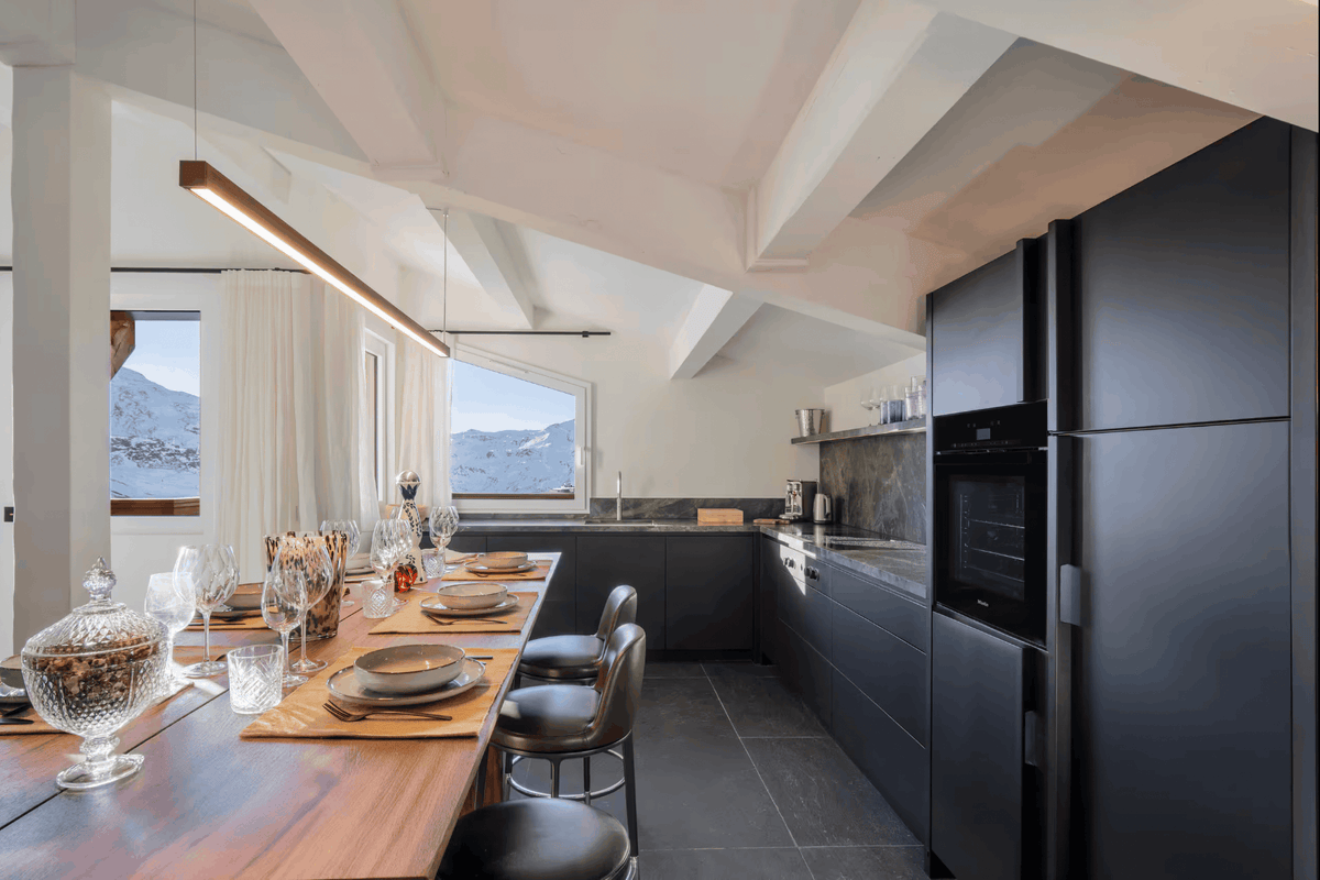 A modern kitchen and dining area with a long wooden table set for dining, dark gray cabinetry, a large black refrigerator, and windows looking out onto a mountain view.