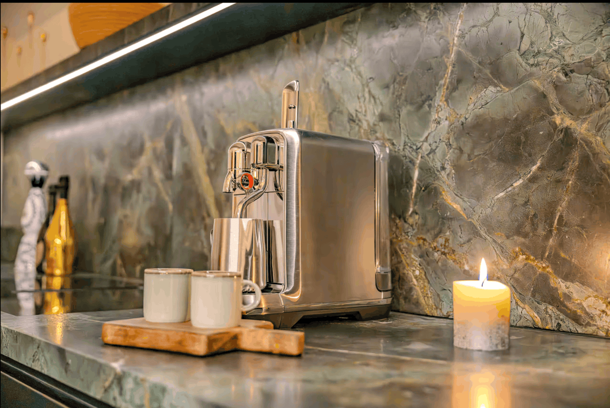 A stainless steel espresso machine sits on a polished stone countertop against a green marble backsplash, with two small white cups on a wooden tray and a lit candle nearby.