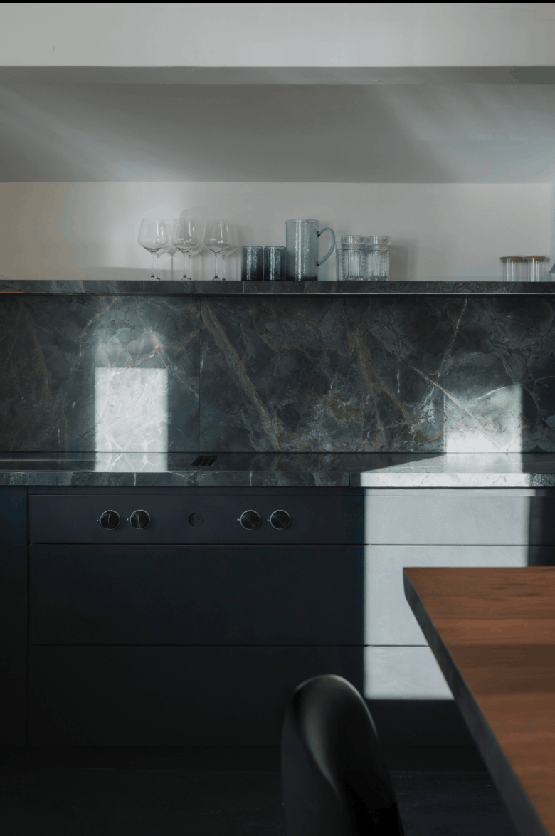 Modern kitchen cabinetry with a dark marble backsplash, a black cooktop, and a wooden table edge in the foreground.