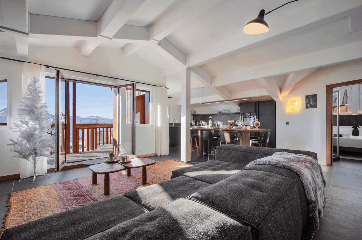 A modern open-plan living space featuring a large dark gray sectional sofa in the foreground, a wooden coffee table, a patterned rug, and large glass doors opening to a balcony with a view of mountains. In the background, a kitchen and dining area are visible.