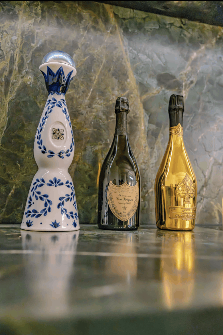 Three decorative bottles lined up on a polished stone surface against a marbled green and gold background. The left bottle is white with blue floral patterns and a blue cap; the middle bottle is dark green with a gold label; the right bottle is metallic gold with a champagne-style label and foil.