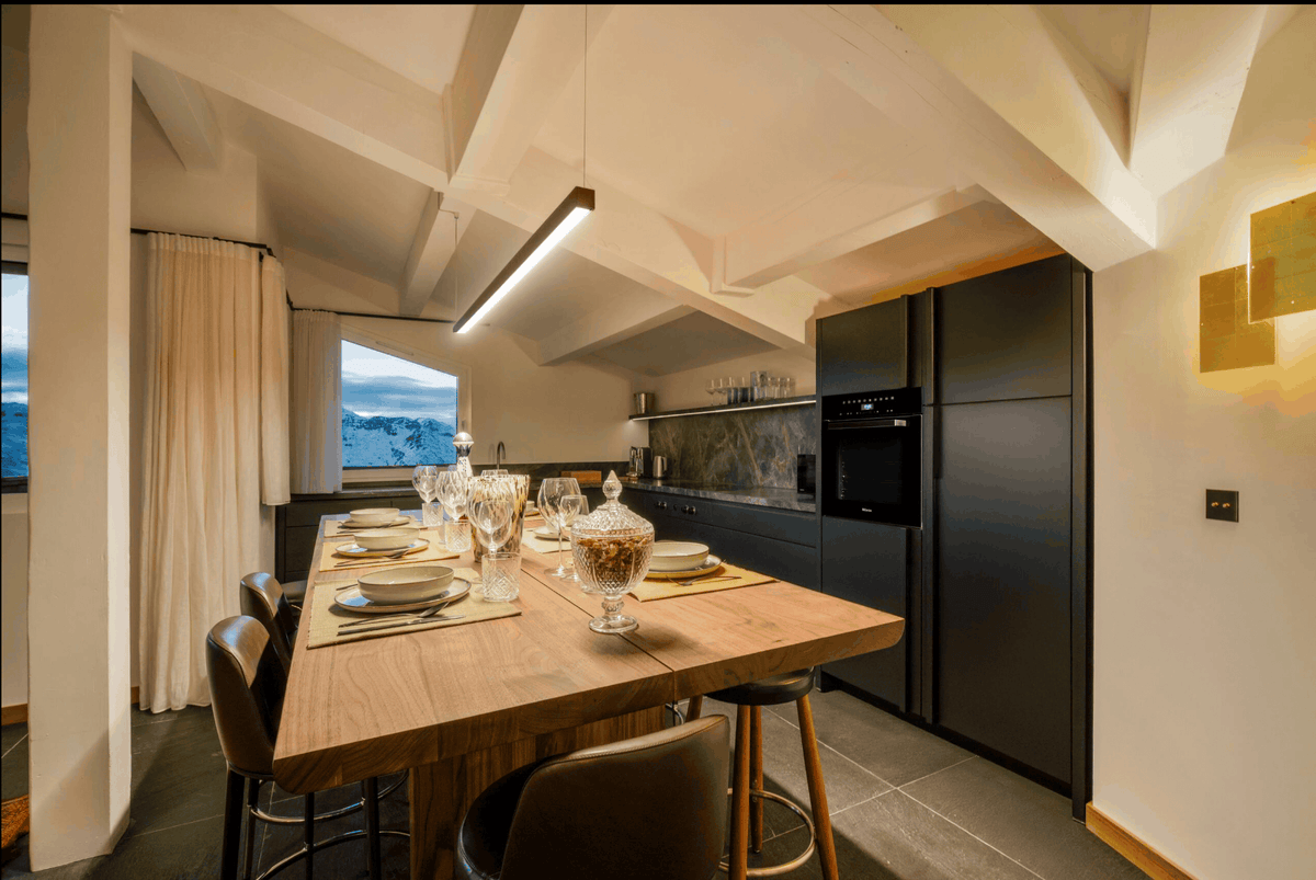 A modern kitchen and dining area with a tall wooden island table set with place settings and bar stools, dark cabinetry, a slanted ceiling with pendant lights, and a window showing an outdoor view.