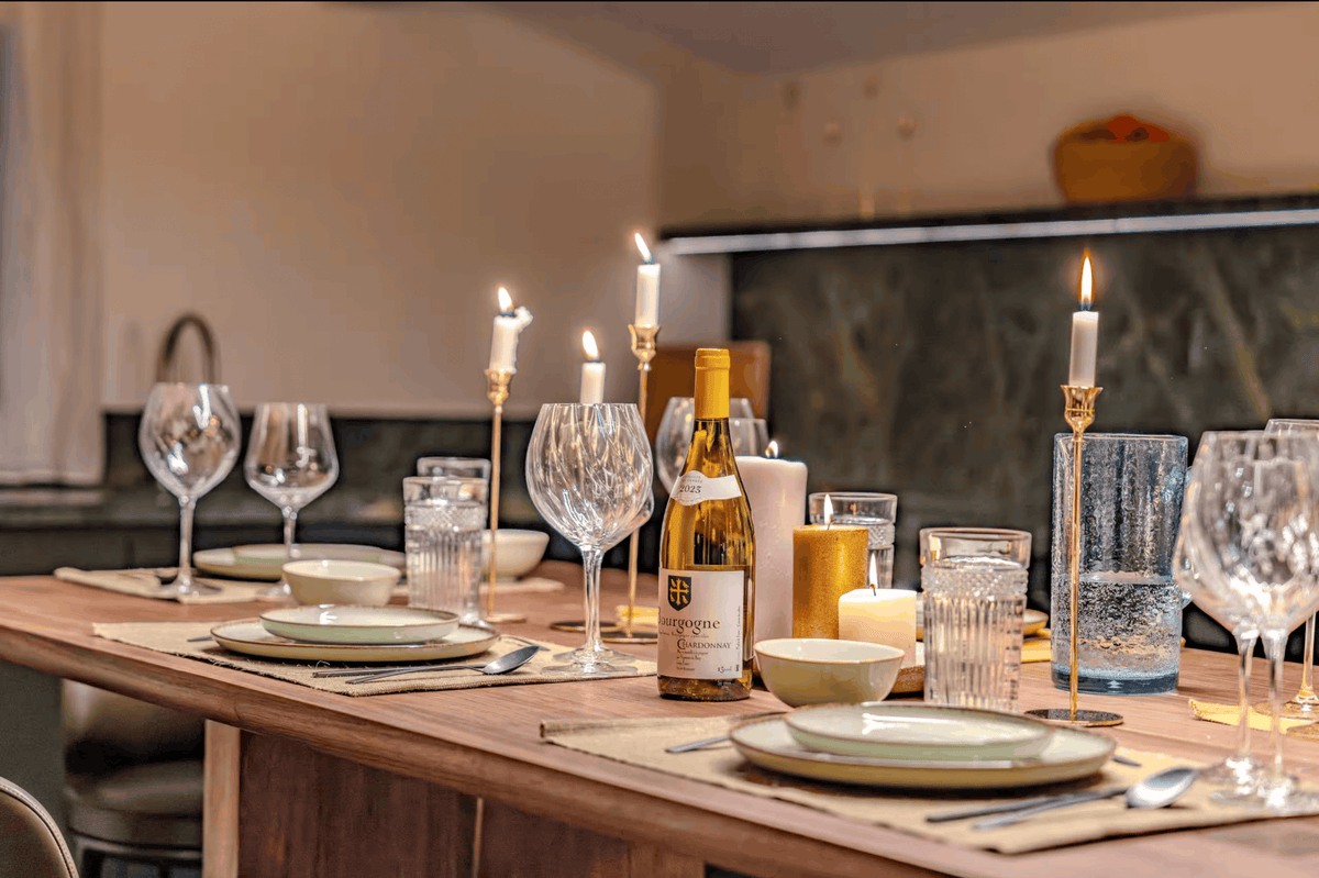 A styled dining table set for guests with wine glasses, plates, candles, and a bottle of wine in a warm, rustic kitchen setting.