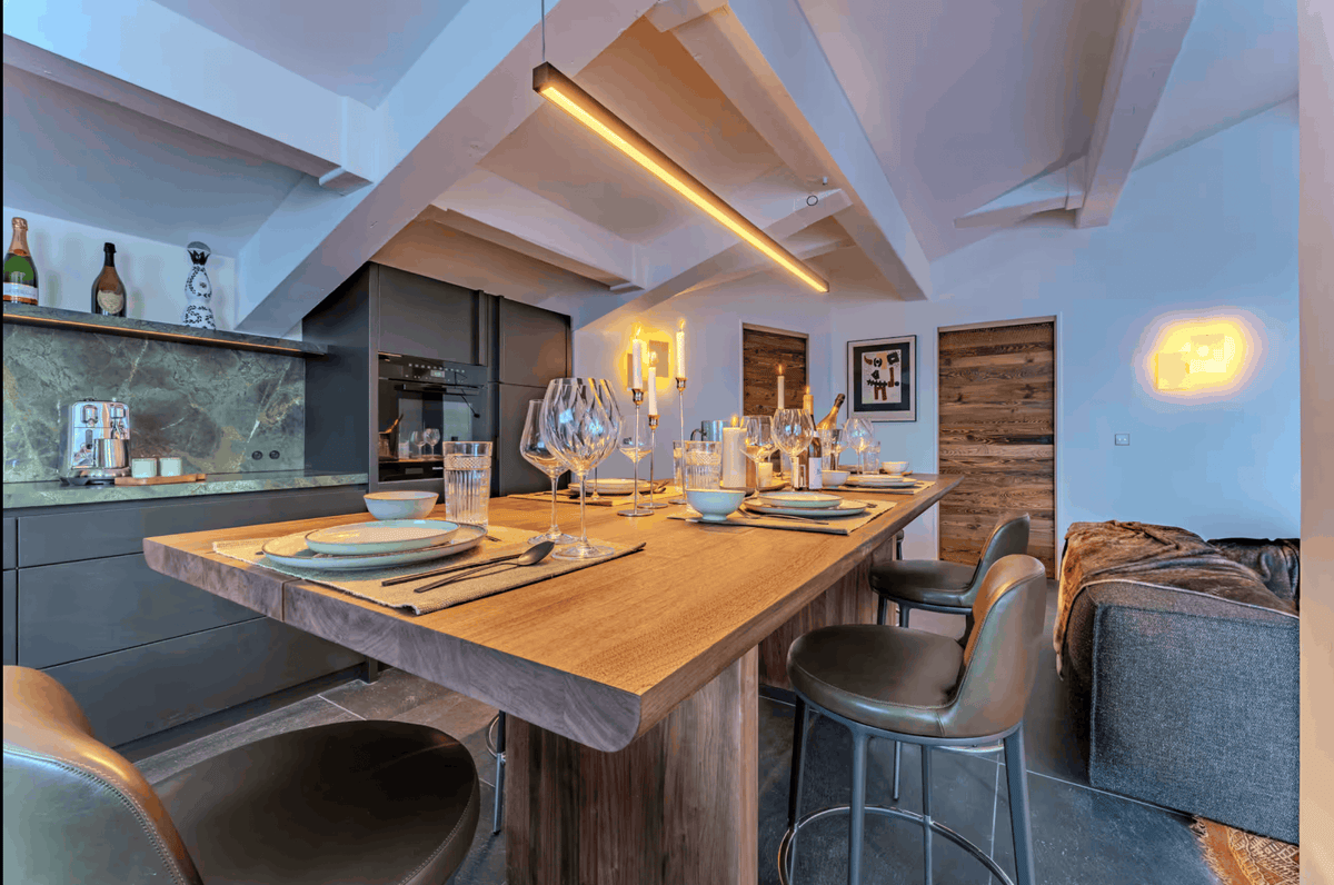 A modern kitchen and dining area with a large wooden bar-height table set with wine glasses and place settings, surrounded by dark bar stools. The space features a slanted ceiling with exposed beams, a built-in kitchen cabinetry unit with a green marble backsplash, and warm ambient lighting.