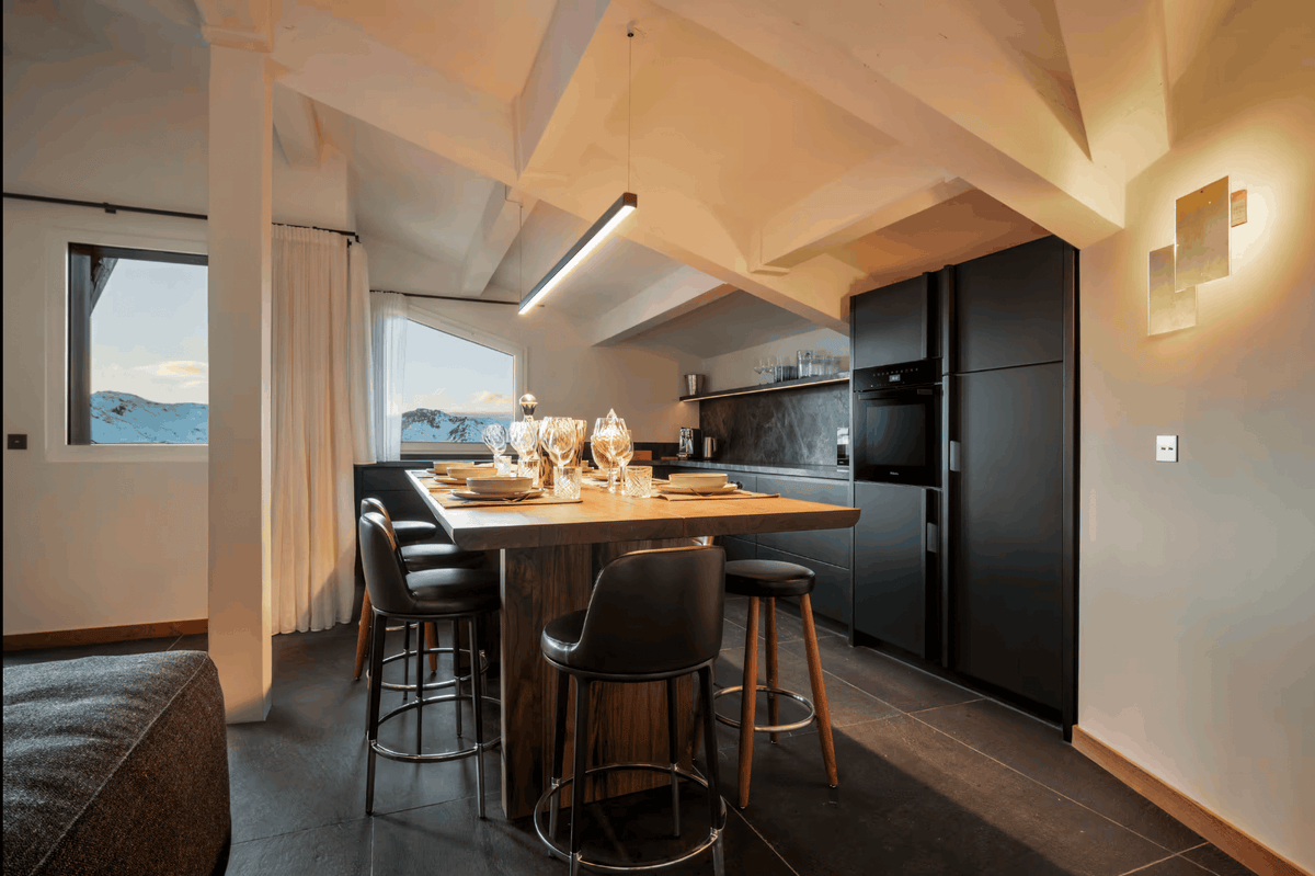 A modern kitchen island with a wooden countertop and black bar stools, set in a spacious kitchen with dark appliances, a high slanted ceiling, and a window showing a mountain view.
