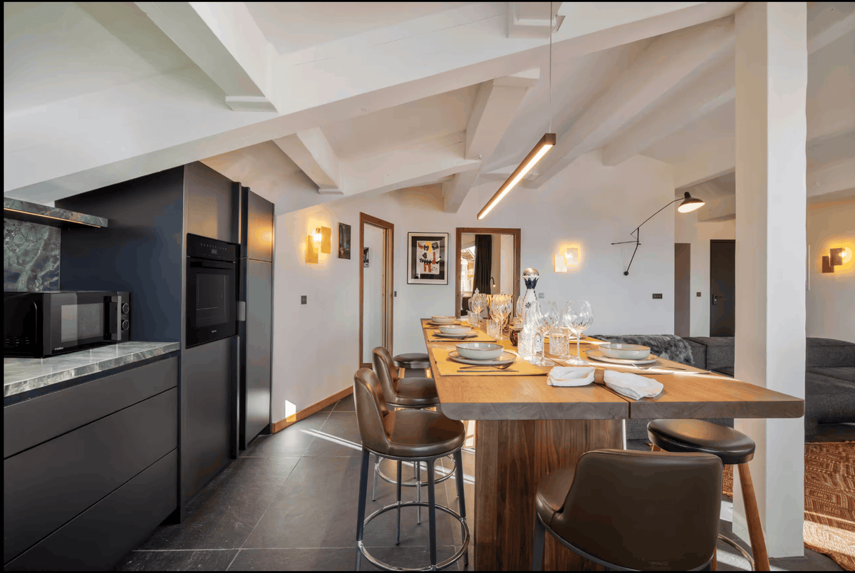 A modern kitchen and dining area with a tall wooden island bar, brown leather bar stools, dark cabinetry, and a bright, airy interior with visible ceiling beams.