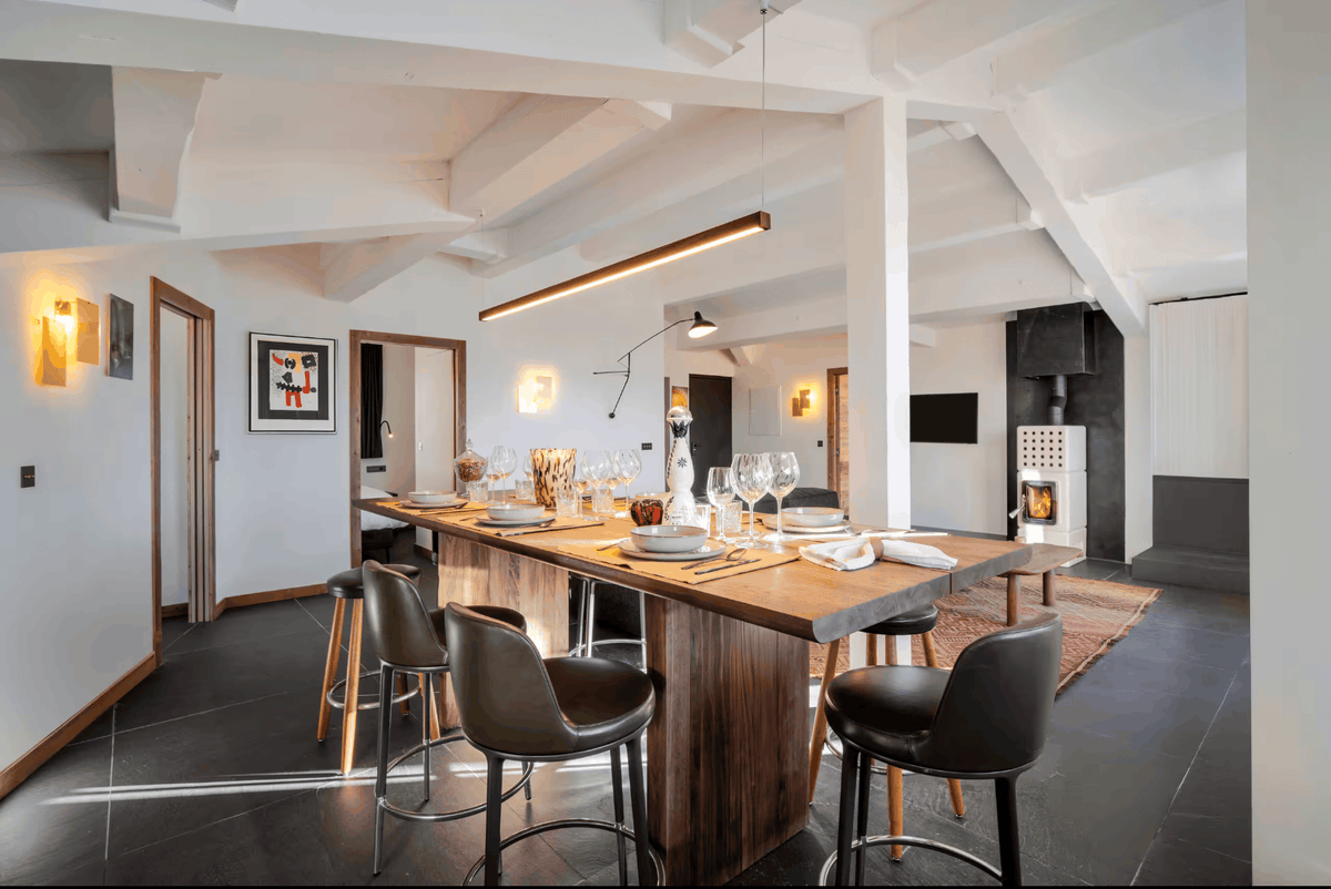 A modern kitchen and dining area featuring a long wooden island table set with glassware, surrounded by four dark leather bar stools. The space has a high vaulted ceiling with white beams, dark gray floor tiles, and a white wood-burning stove in the background.
