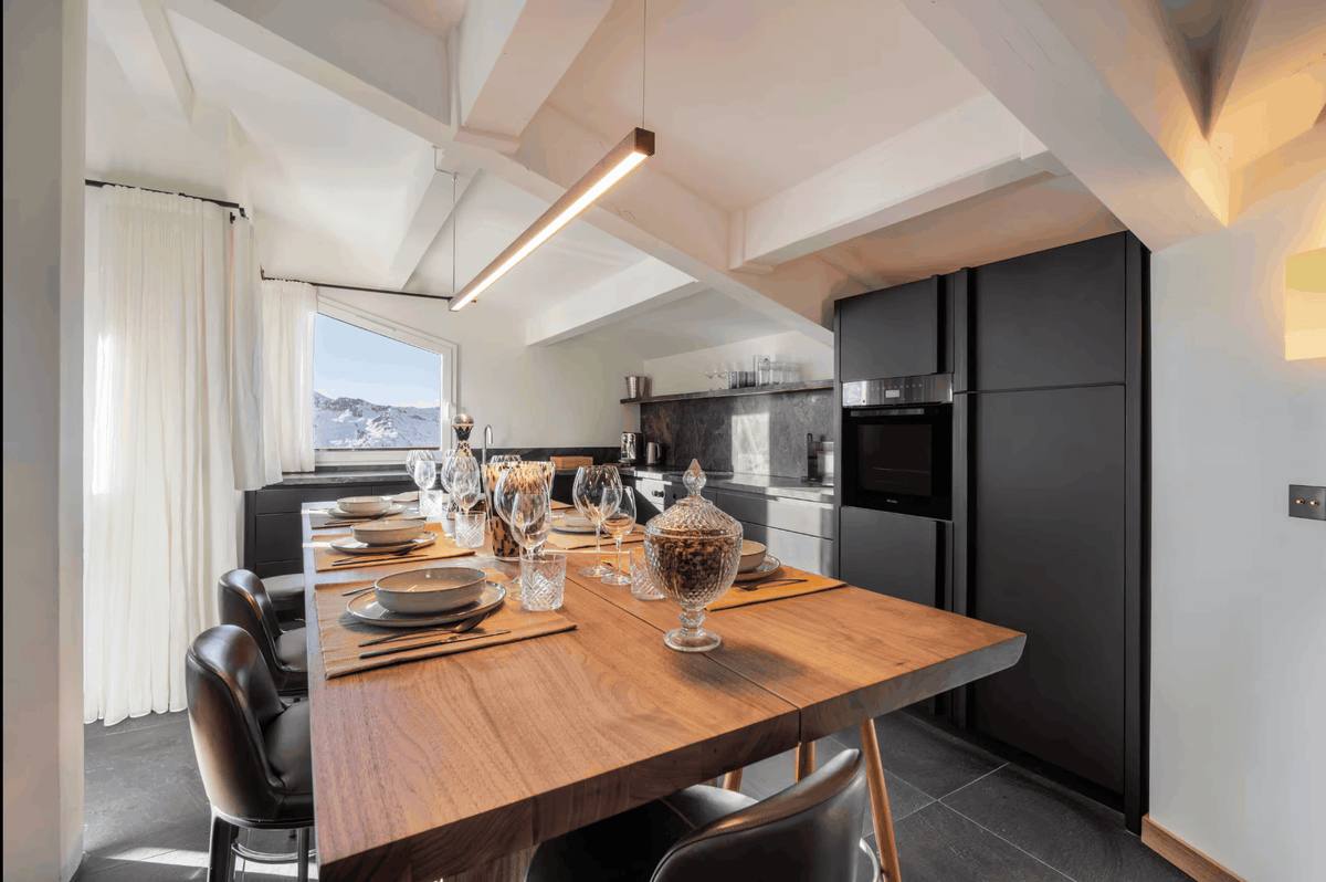 A modern kitchen and dining area with a large wooden dining table set with plates and glasses, black kitchen cabinets, a window with white curtains, and a slanted ceiling with exposed beams.