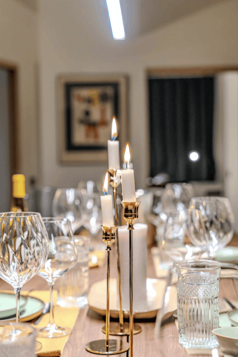 A dining table setting with lit white taper candles in gold holders, surrounded by clear glass drinking glasses and plates in a softly blurred indoor room.