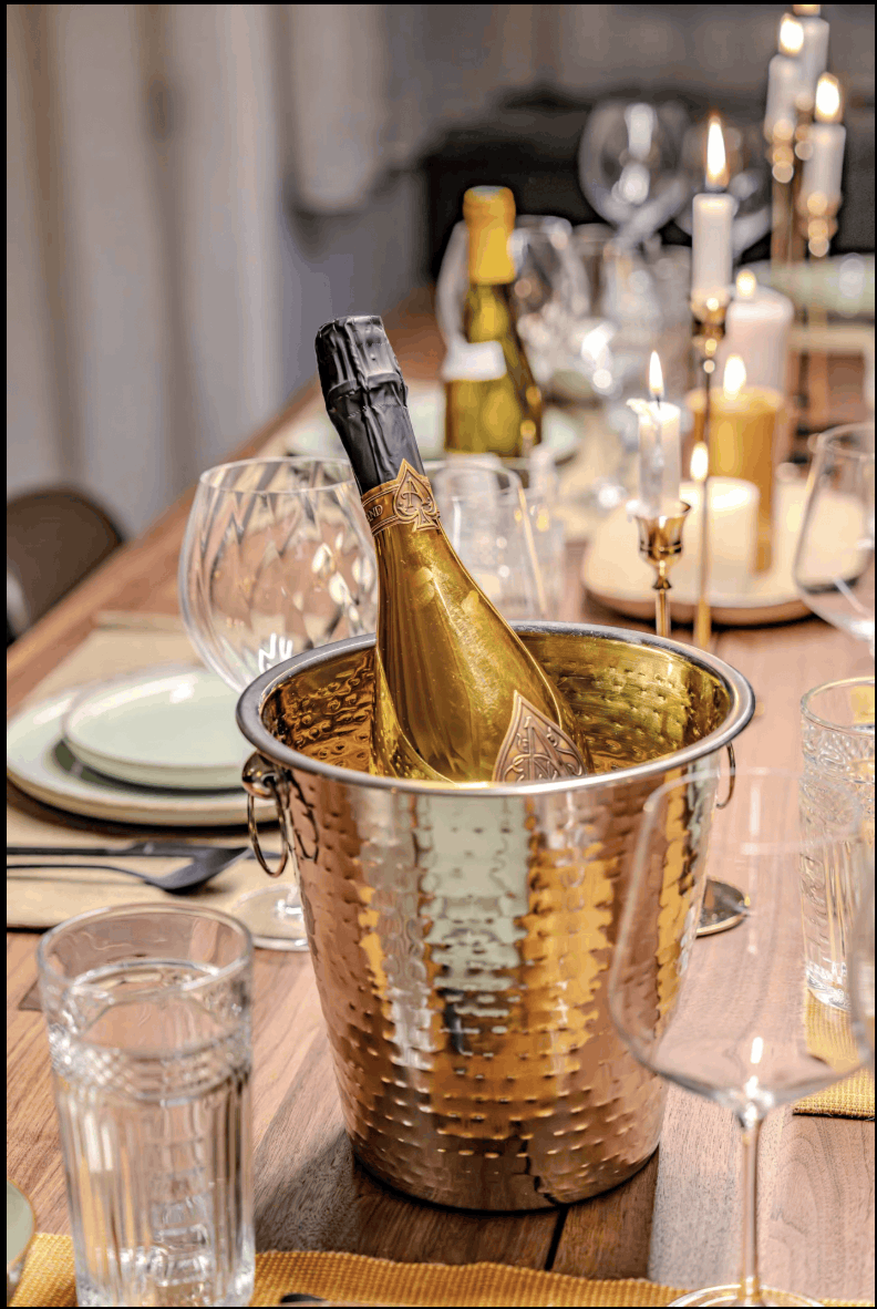 A hammered copper champagne bucket holding a gold-toned champagne bottle, set on a wooden dining table surrounded by glassware, plates, and lit candles.