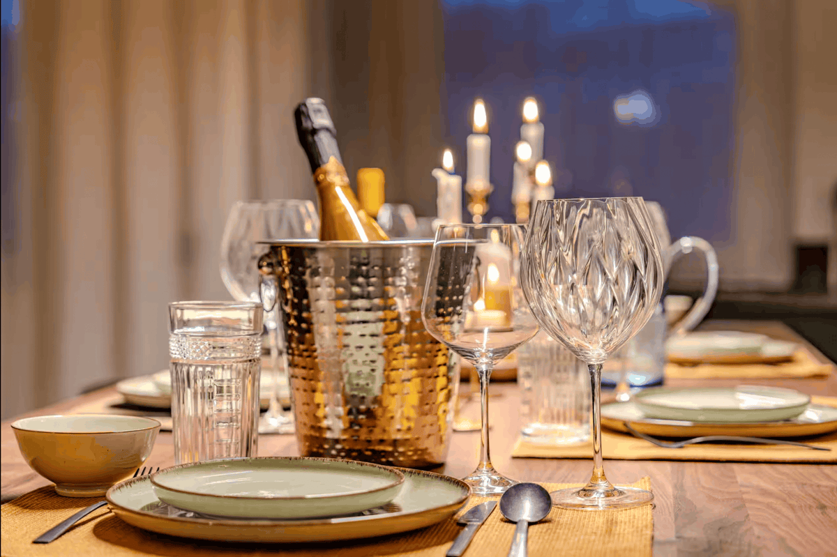 A styled dining table setting with a hammered metal champagne bucket holding a bottle, clear cut wine glasses, water glasses, plates, and candles in the background.