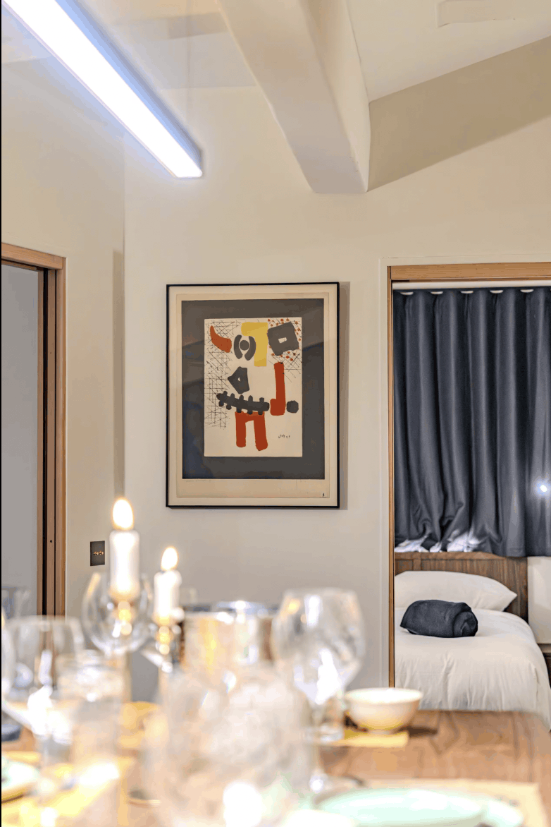 A framed abstract art print with bold red, black, yellow, and gray shapes hangs on a beige wall in a modern bedroom setting. The room features a bed with white linens and a dark gray curtain, with a blurred dining table and glassware in the foreground.