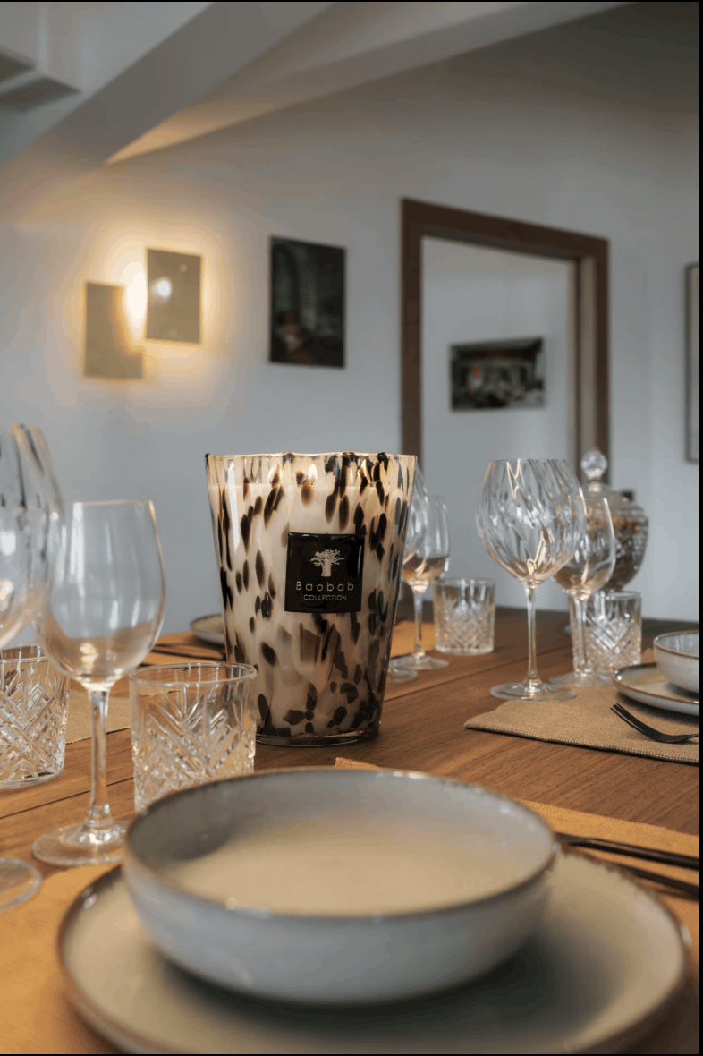 A tall glass candle holder with a black and white leopard print pattern, featuring a central black label with a white bull logo and the brand name 'BULLION'. It is styled on a wooden dining table surrounded by wine glasses, crystal tumblers, and ceramic plates in a warmly lit home interior.