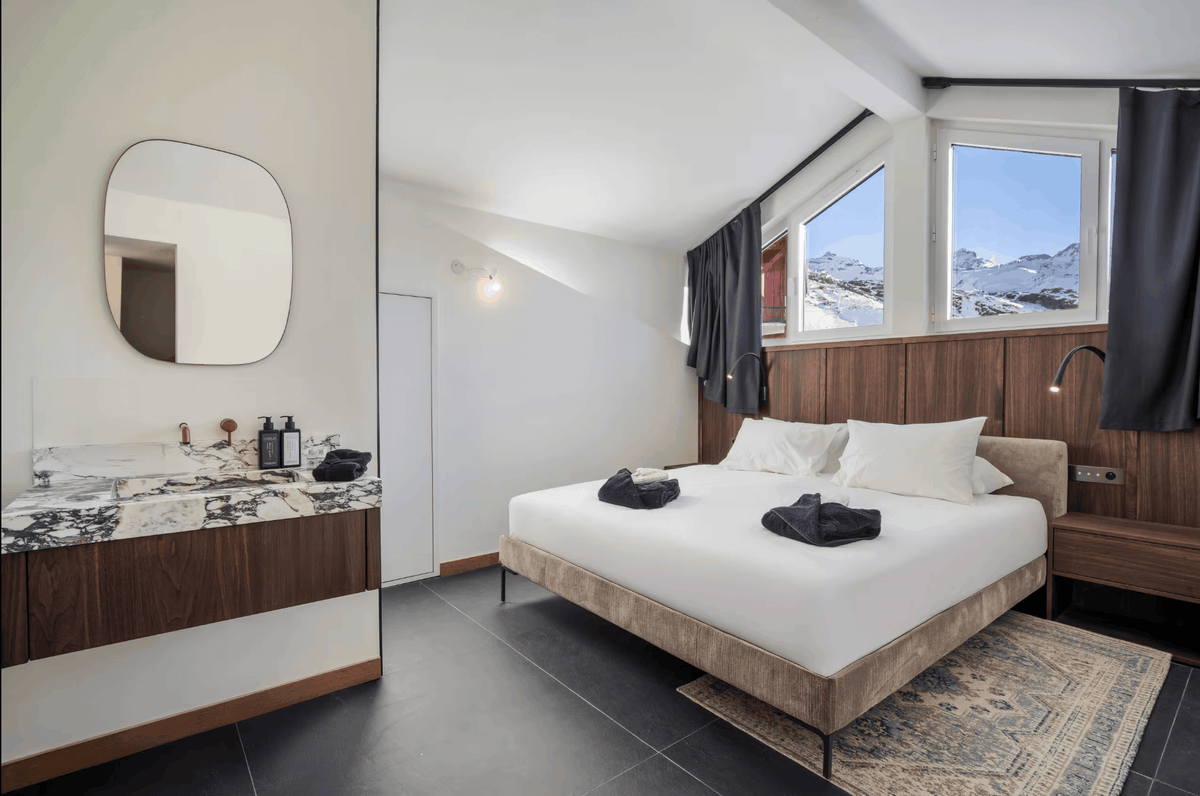 A modern ski resort bedroom featuring a double bed with white bedding, a wood-paneled headboard, large windows with a view of snowy mountains, a vanity with a marble countertop, and a patterned rug.