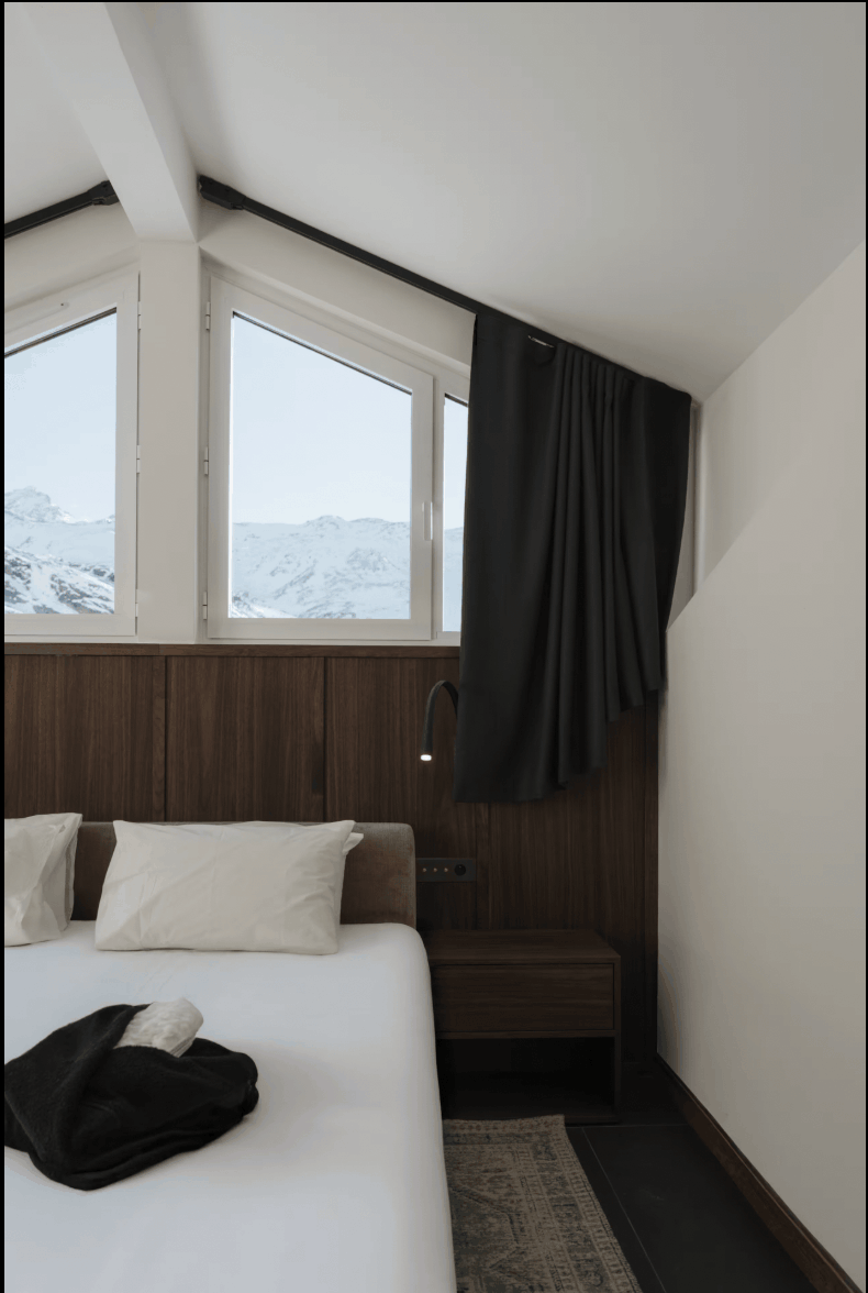 A modern bedroom interior with a bed dressed in white bedding and a black folded garment, set against a wood-paneled wall and large windows showing a snowy mountain view. A black curtain hangs beside the window.