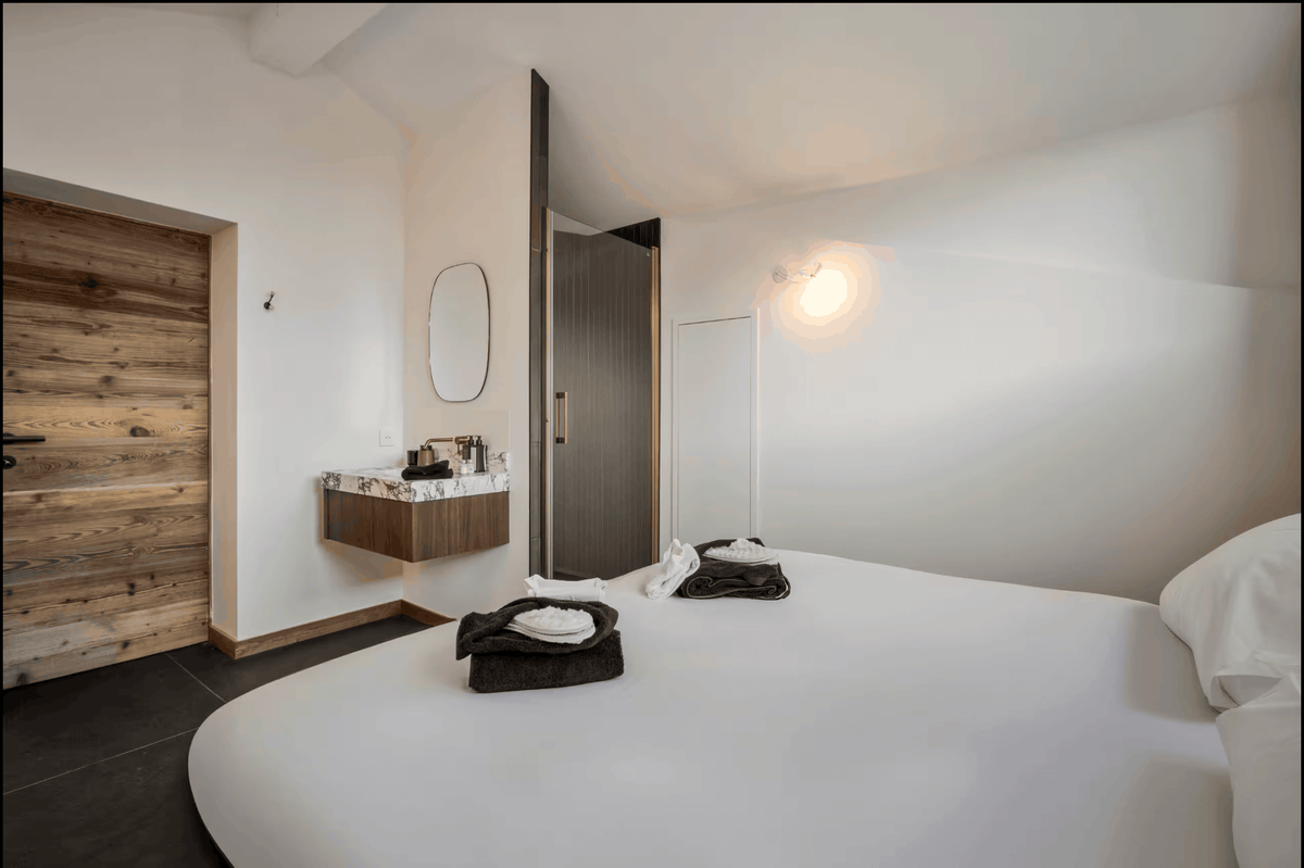 A modern bedroom interior featuring a large bed with white bedding, a floating wooden vanity with a marble sink, a dark wood door, and a warm wall-mounted light.
