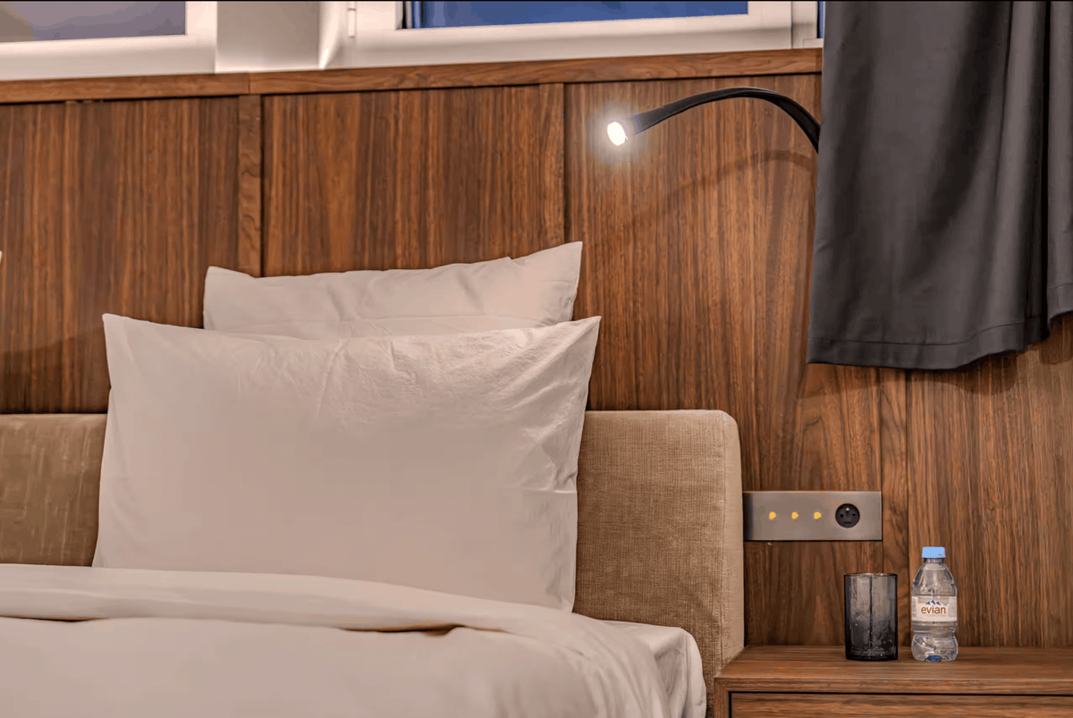 A close-up of a neatly made bed with white pillows and duvet, set against a dark wood-paneled headboard. A black adjustable reading light is mounted above, and a bedside table at right holds a water bottle and a dark glass cup.