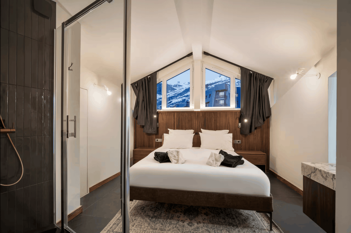 A modern mountain-style bedroom with a large bed, white bedding, dark wood headboard and footboard, and tall arched windows showing snow-capped mountains outside. A glass shower enclosure is visible on the left.