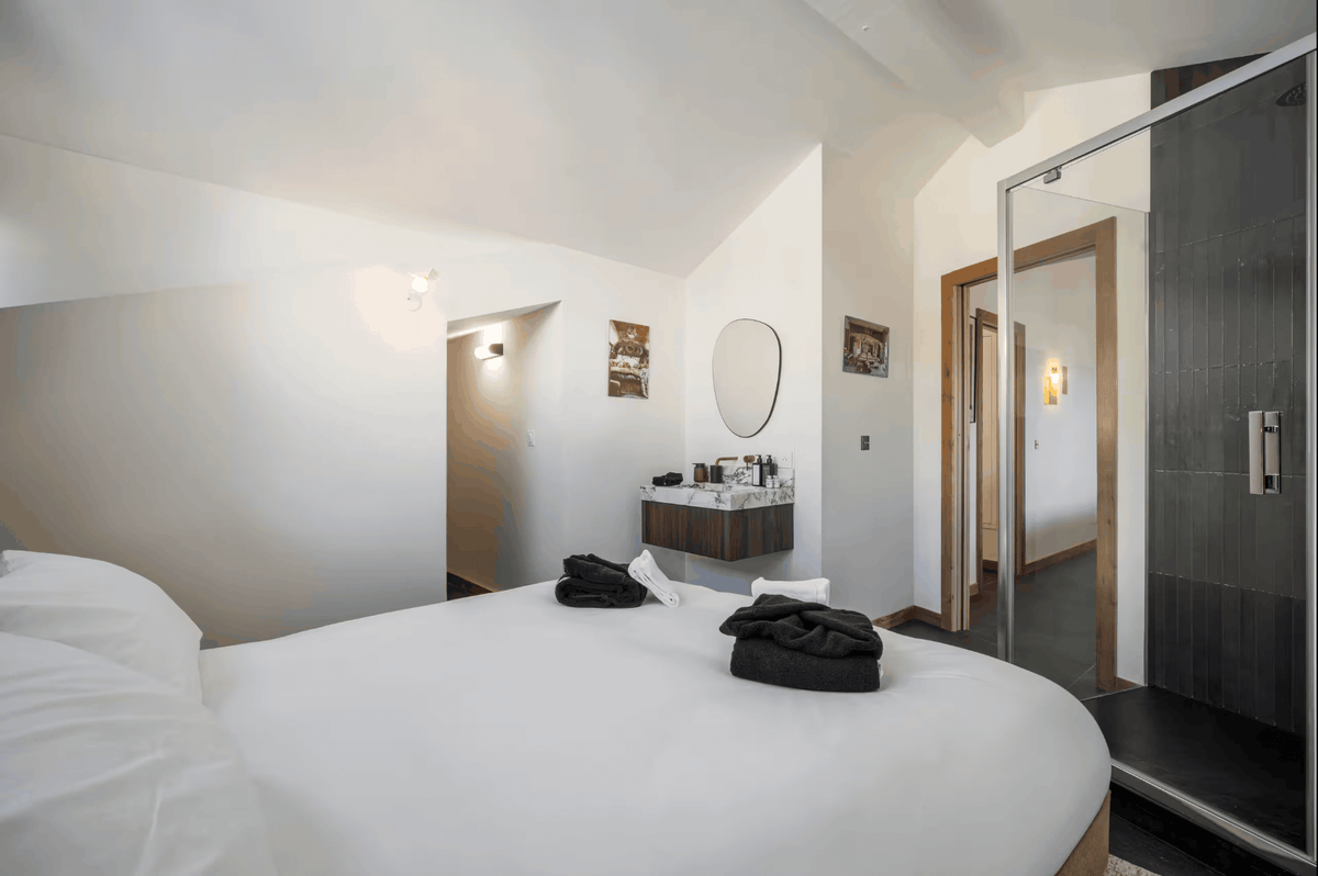 A modern bedroom interior featuring a large bed with white bedding and black folded towels, a small vanity with a round mirror, and a glass shower enclosure on the right.