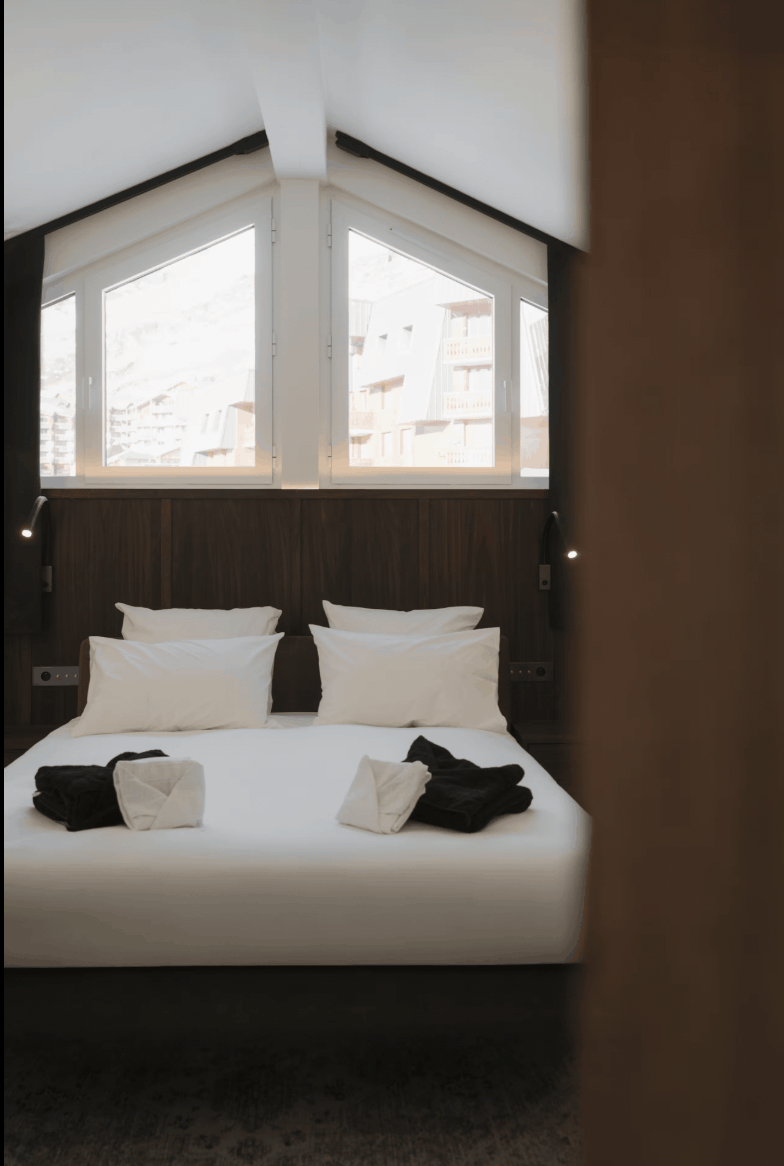 A neatly made bed with white linens and pillows in a modern bedroom, viewed through a dark wooden door frame. The room features a dark wood headboard, built-in wall lights, and large angled windows with a view of buildings outside.