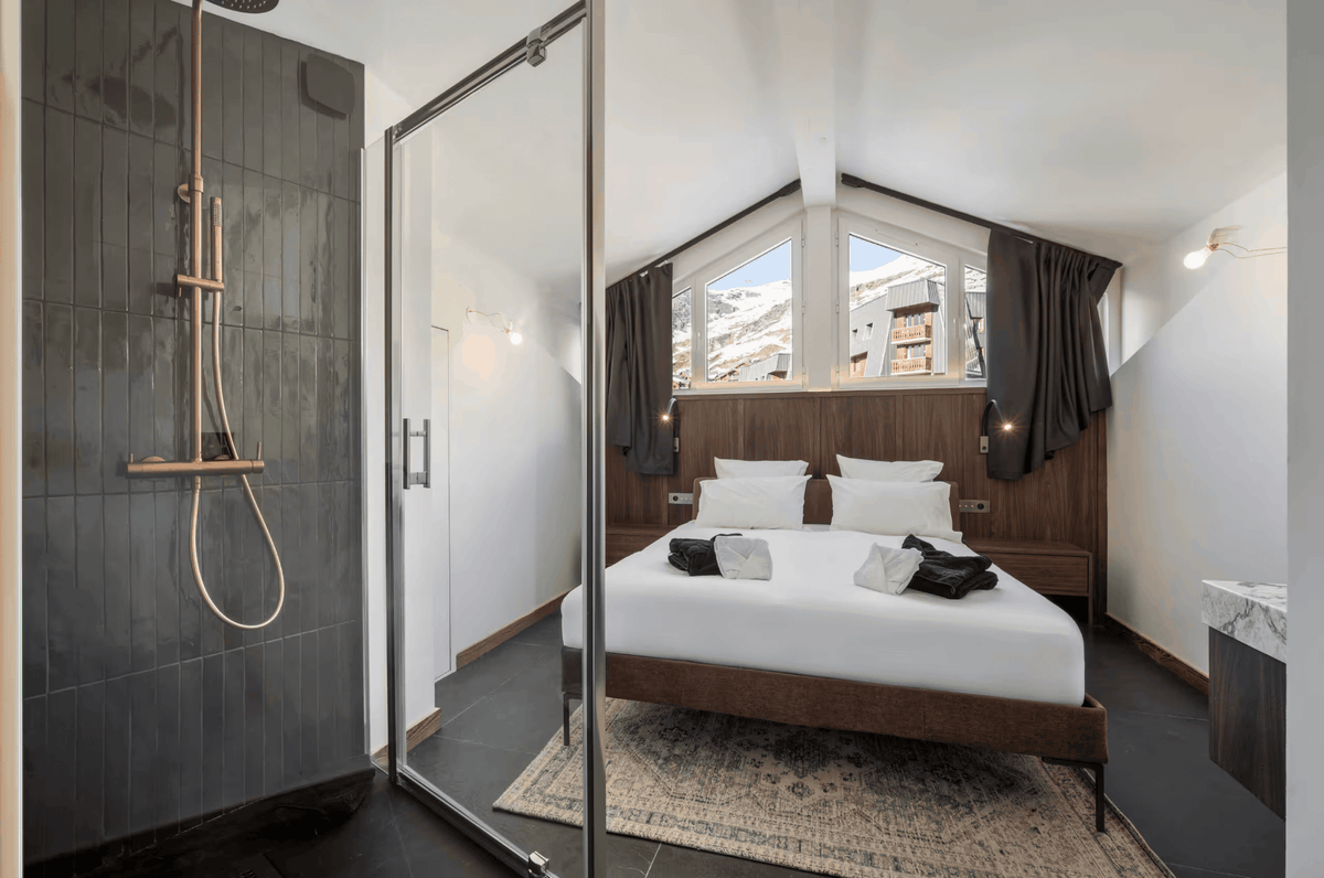 Modern minimalist bedroom with a large bed dressed in white linens, dark wood headboard and footboard, and a glass sliding door leading to a dark gray tiled shower area.