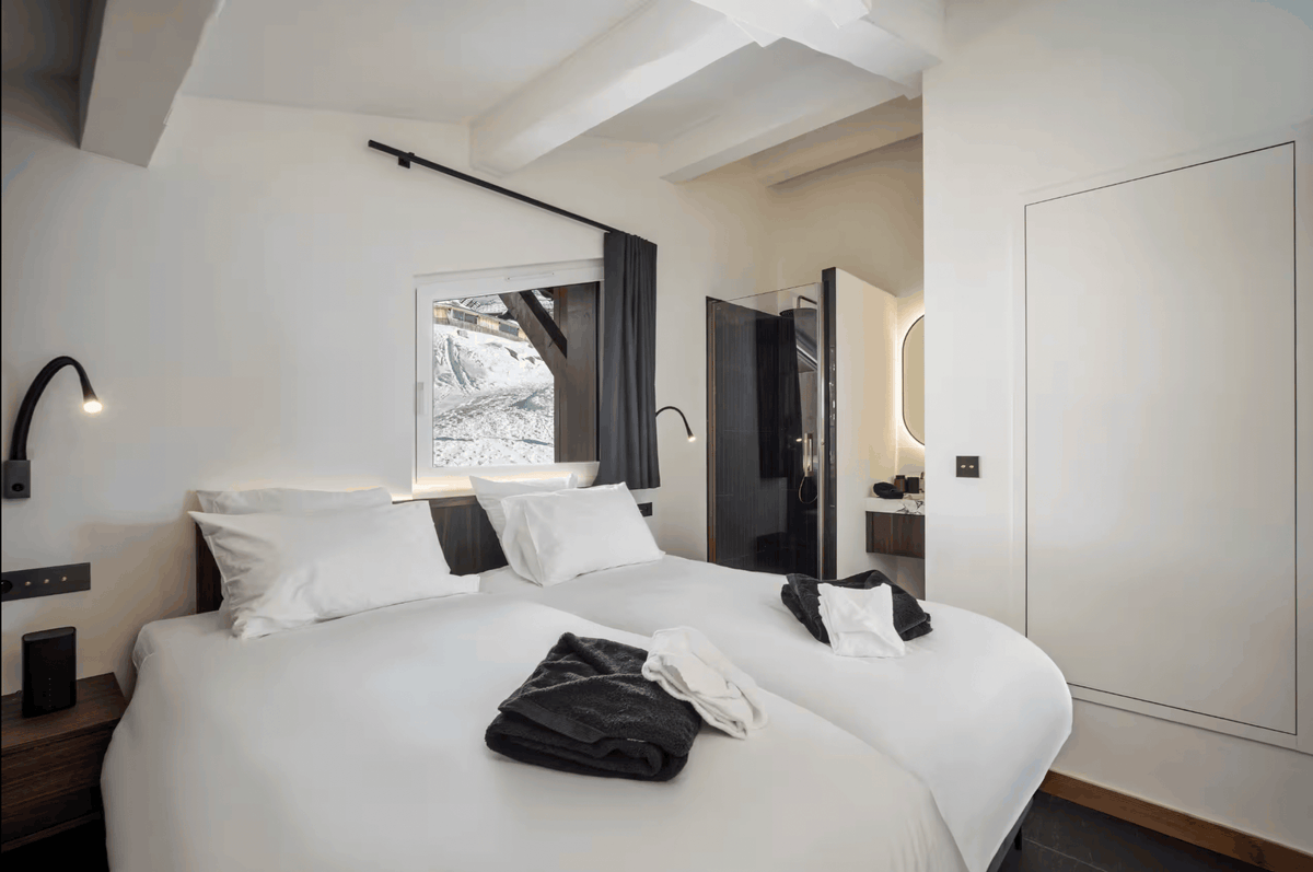 A modern minimalist bedroom featuring a large bed with crisp white linens, black wall-mounted lamps, and a window looking out onto a snowy mountain scene. The room has clean white walls, a built-in wardrobe, and a glimpse of a bathroom in the background.