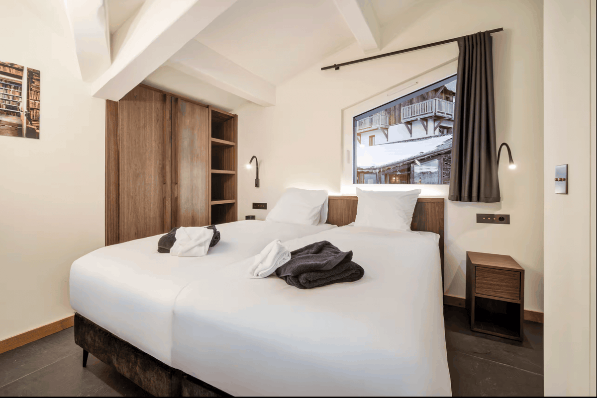 A modern bedroom interior featuring a neatly made bed with white linens, a large window with a view of snowy mountainside, dark gray curtains, wooden furniture, and a warm off-white wall.