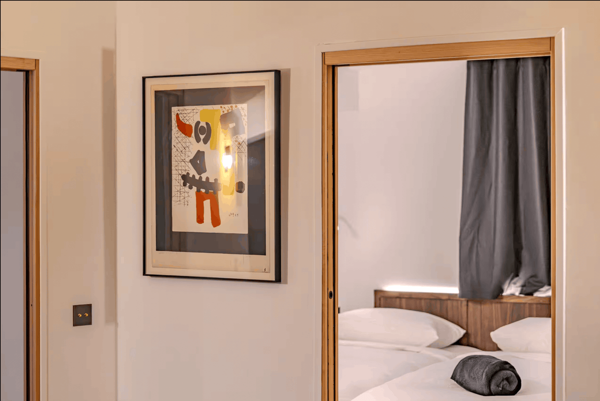 A framed abstract art print hangs on a beige wall in a modern bedroom setting, with a mirror reflecting a bed with white linens and a gray curtain.