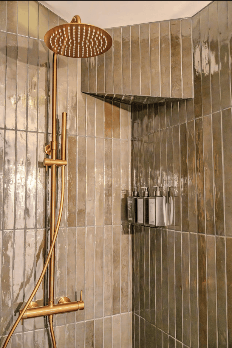 A modern shower corner with glossy vertical stone tiles in warm gray-brown tones and a brushed gold shower fixture including a round overhead showerhead, handheld wand, and control bar. Small silver toiletry bottles are mounted on the tiled wall.