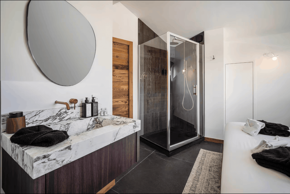 Modern bathroom interior with a white marble sink and countertop, dark wood vanity, round wall mirror, glass shower enclosure, and a white bed with black towels in the room.