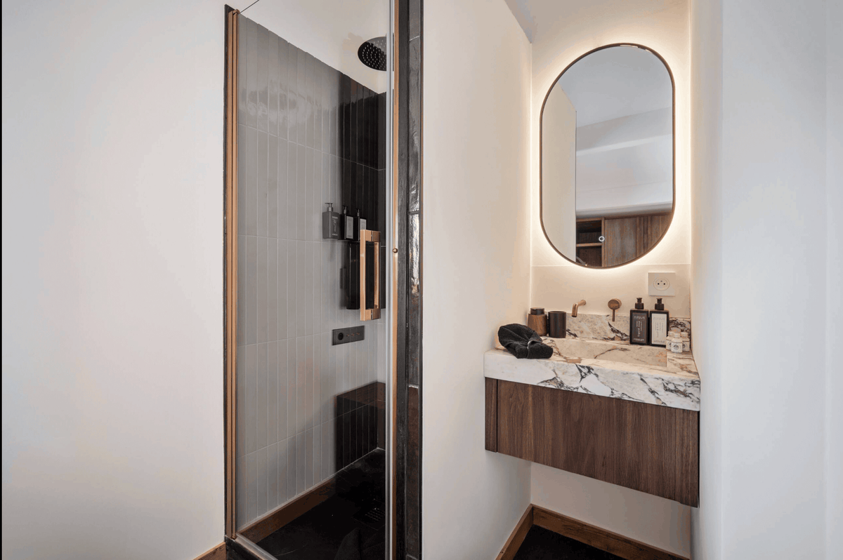 Modern bathroom interior featuring a walk-in shower with a glass door and dark wood trim, alongside a floating vanity with a white marble countertop, dark wood cabinet, and an oval backlit mirror.