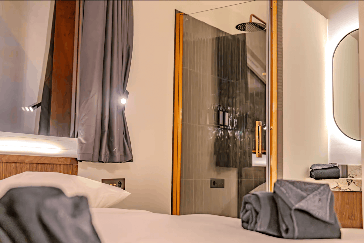 A modern hotel room interior featuring a neatly made bed with white linens and a folded gray towel in the foreground, an open glass shower door with warm wood framing, and a softly lit bathroom area.