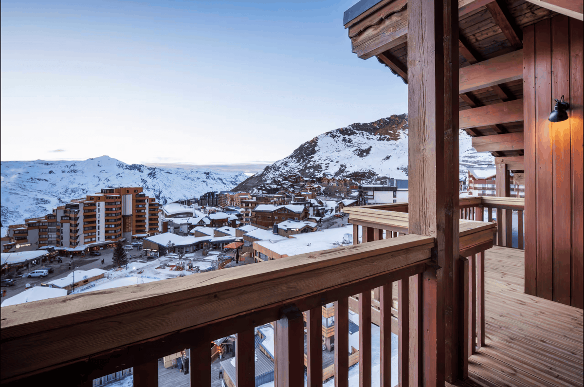 A scenic view from a wooden balcony overlooking a snowy mountain village under a clear blue sky, with snow-covered peaks and resort buildings in the distance.
