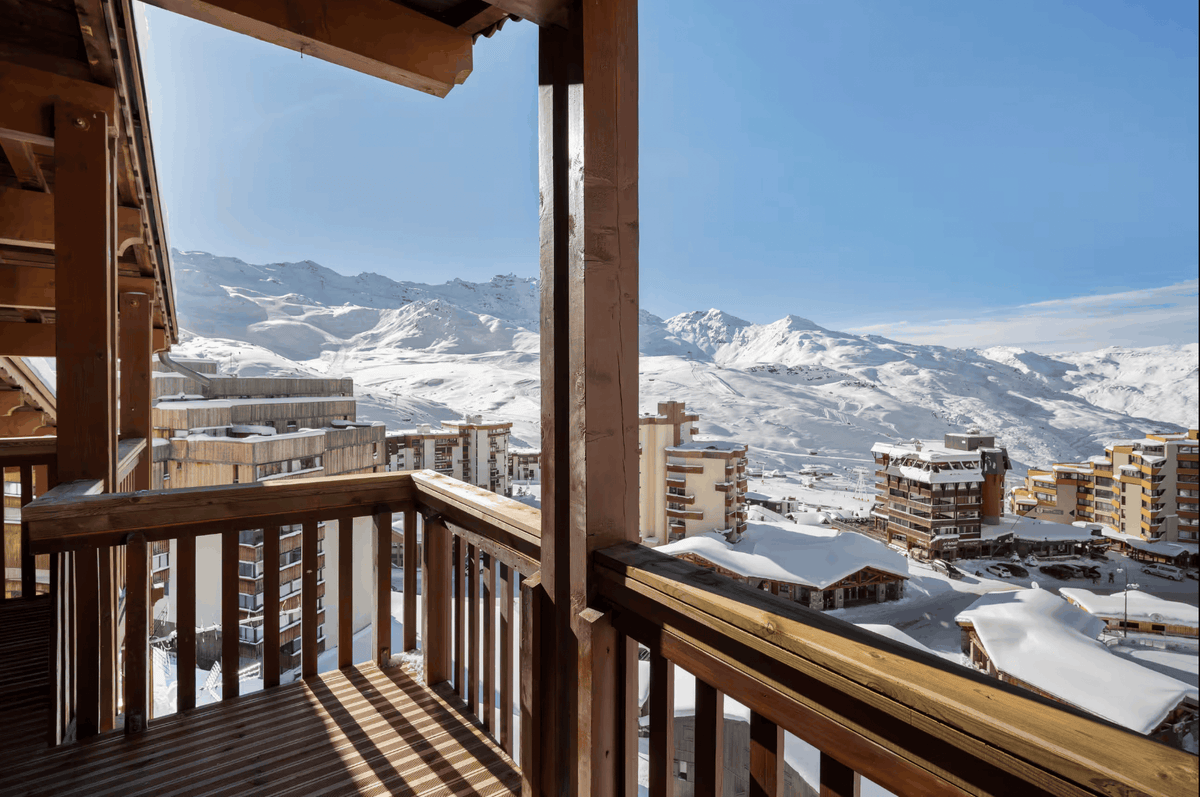 A wooden balcony overlooking a snowy mountain village under a clear blue sky, with snow-covered peaks and resort buildings in the background.