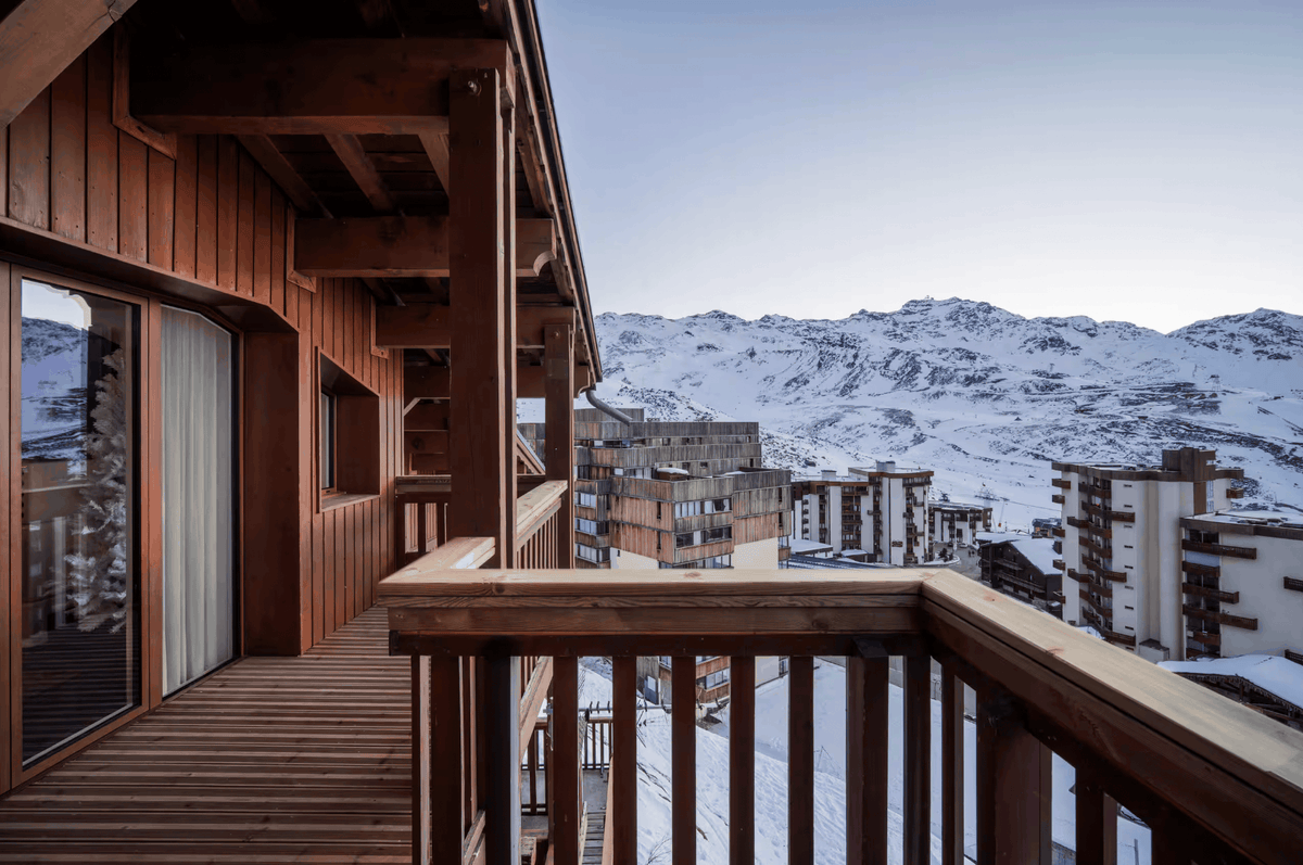A wooden balcony overlooking a snowy mountain village with snow-covered peaks under a pale blue sky.