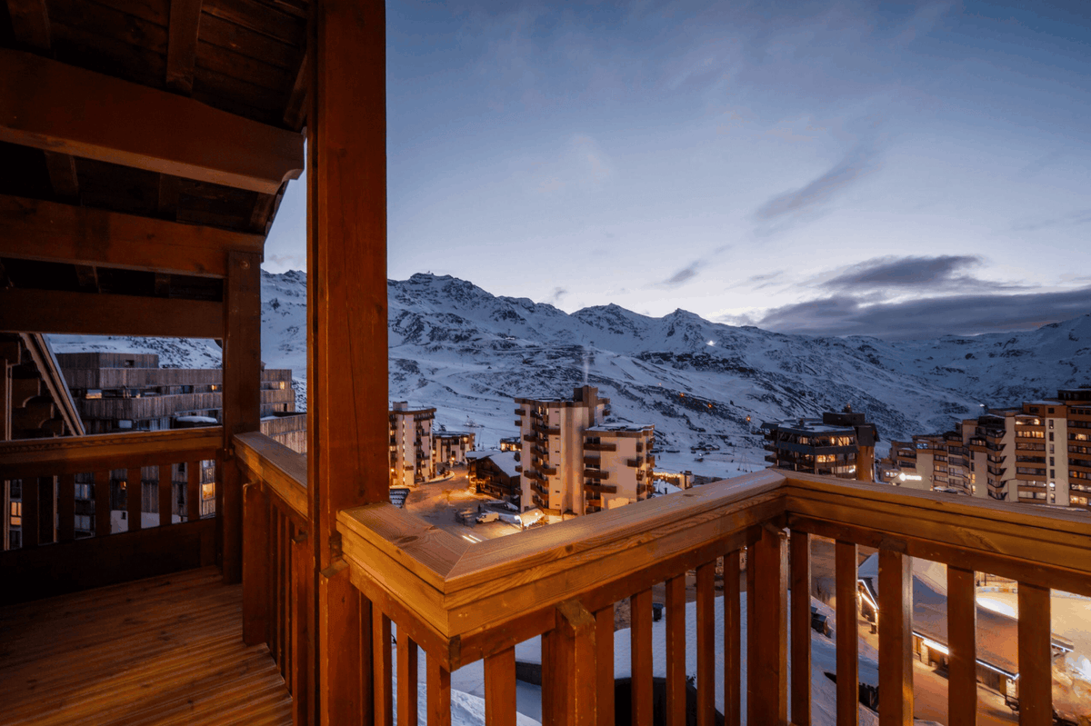 A warm wooden balcony overlooking a snowy mountain village at dusk, with snow-covered peaks and blue evening sky in the background.