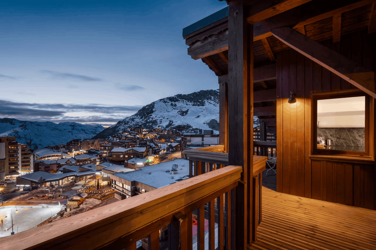 A warm wooden chalet balcony overlooking a snowy mountain village at dusk, with a blue twilight sky and illuminated buildings in the valley below.