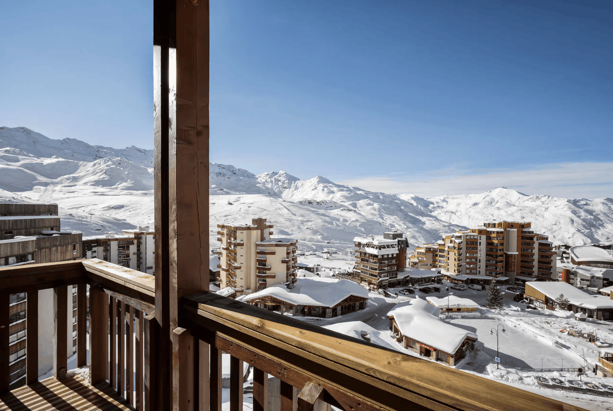 A scenic view from a wooden balcony overlooking a snowy mountain village with chalets and snow-covered peaks under a clear blue sky.