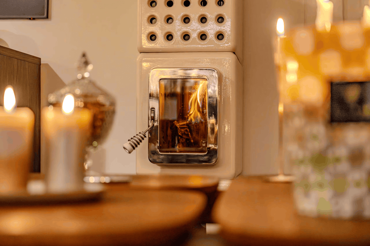 A cream-colored wall-mounted wood-burning stove with a glass front door showing a fire, surrounded by warm, softly blurred candles and wooden surfaces in a cozy interior setting.