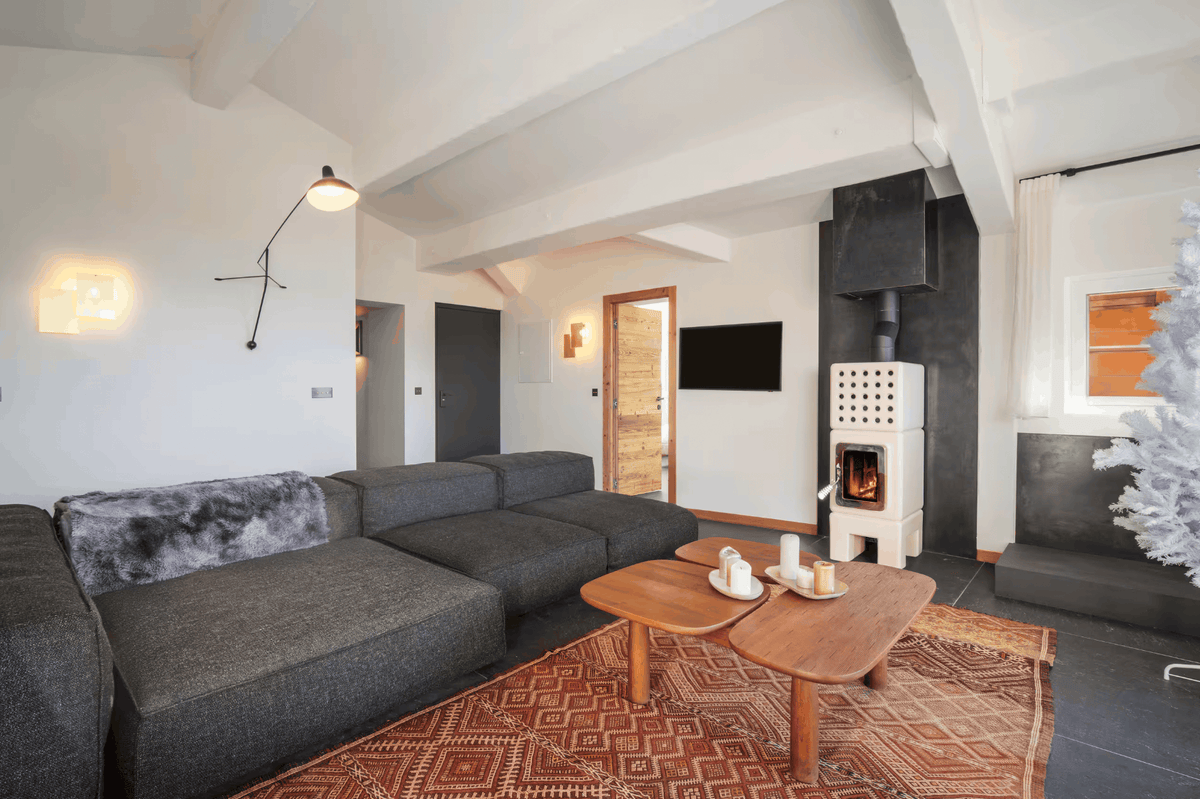 A modern living room featuring a dark gray sectional sofa, two wooden coffee tables, a patterned terracotta rug, and a white wood-burning stove against a dark wall.